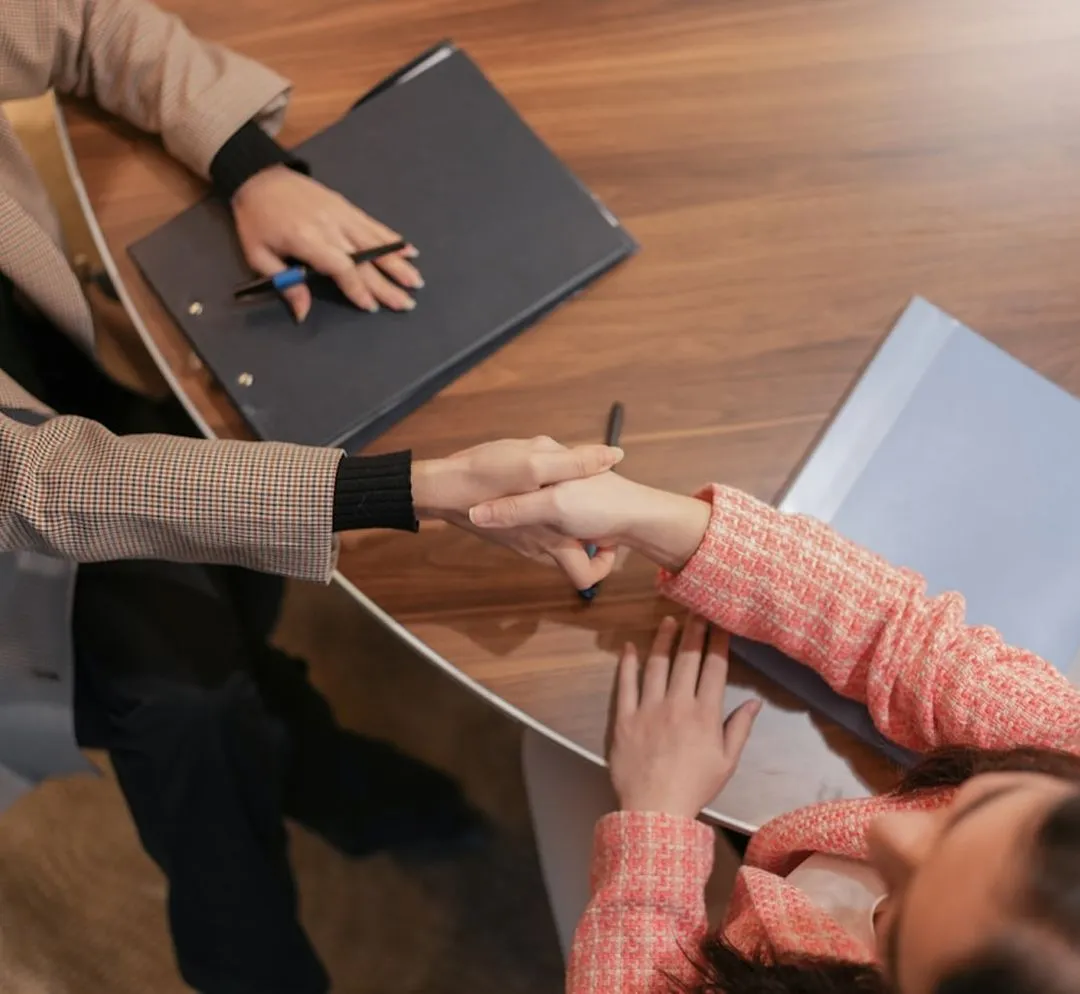 Deux personnes échangeant une poignée de main par-dessus une table, avec des documents et un stylo à proximité.