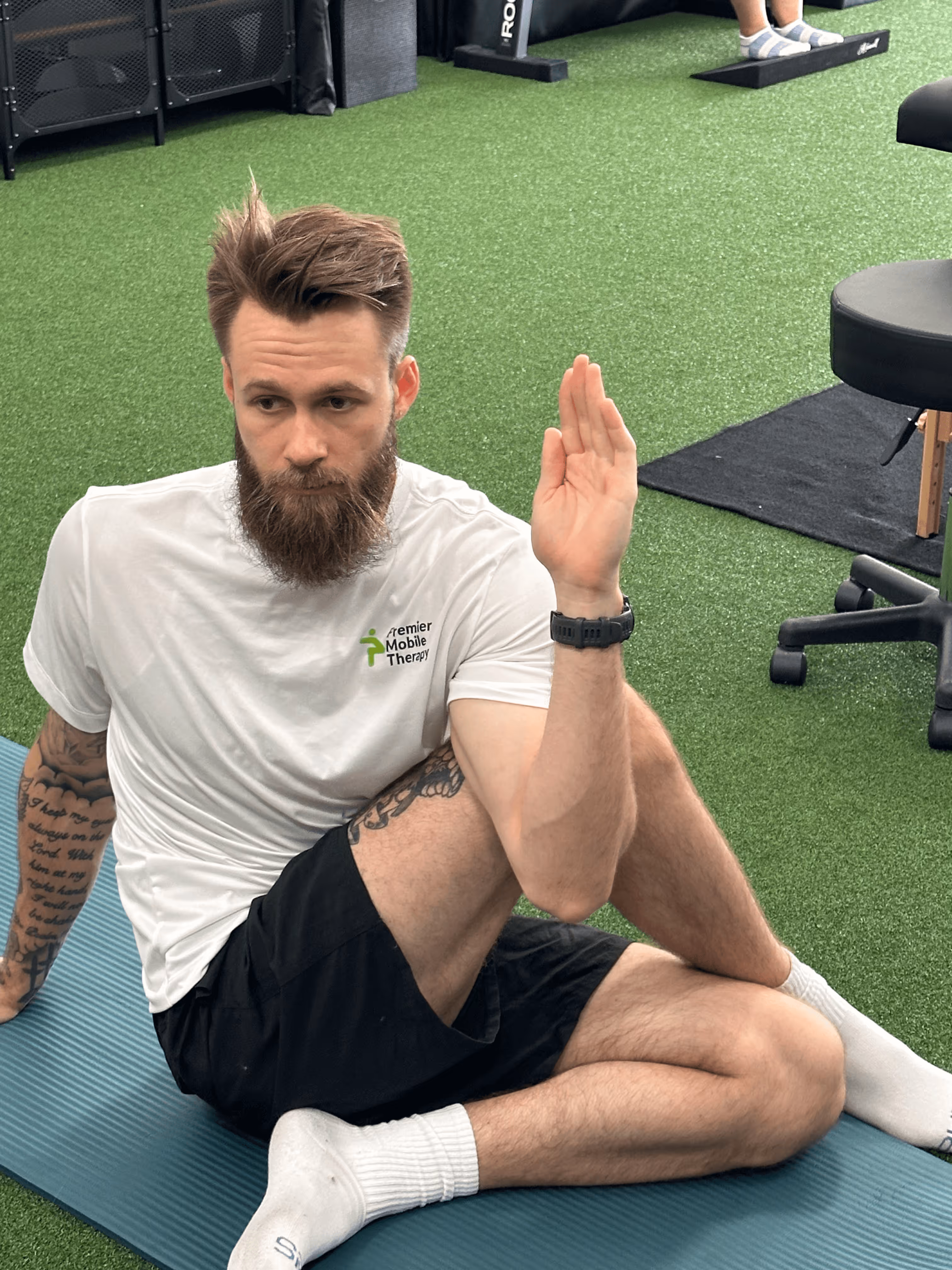 Bearded man in a white Premier Mobile Therapy shirt performing a seated spinal twist stretch on a blue mat in a gym with artificial green turf.