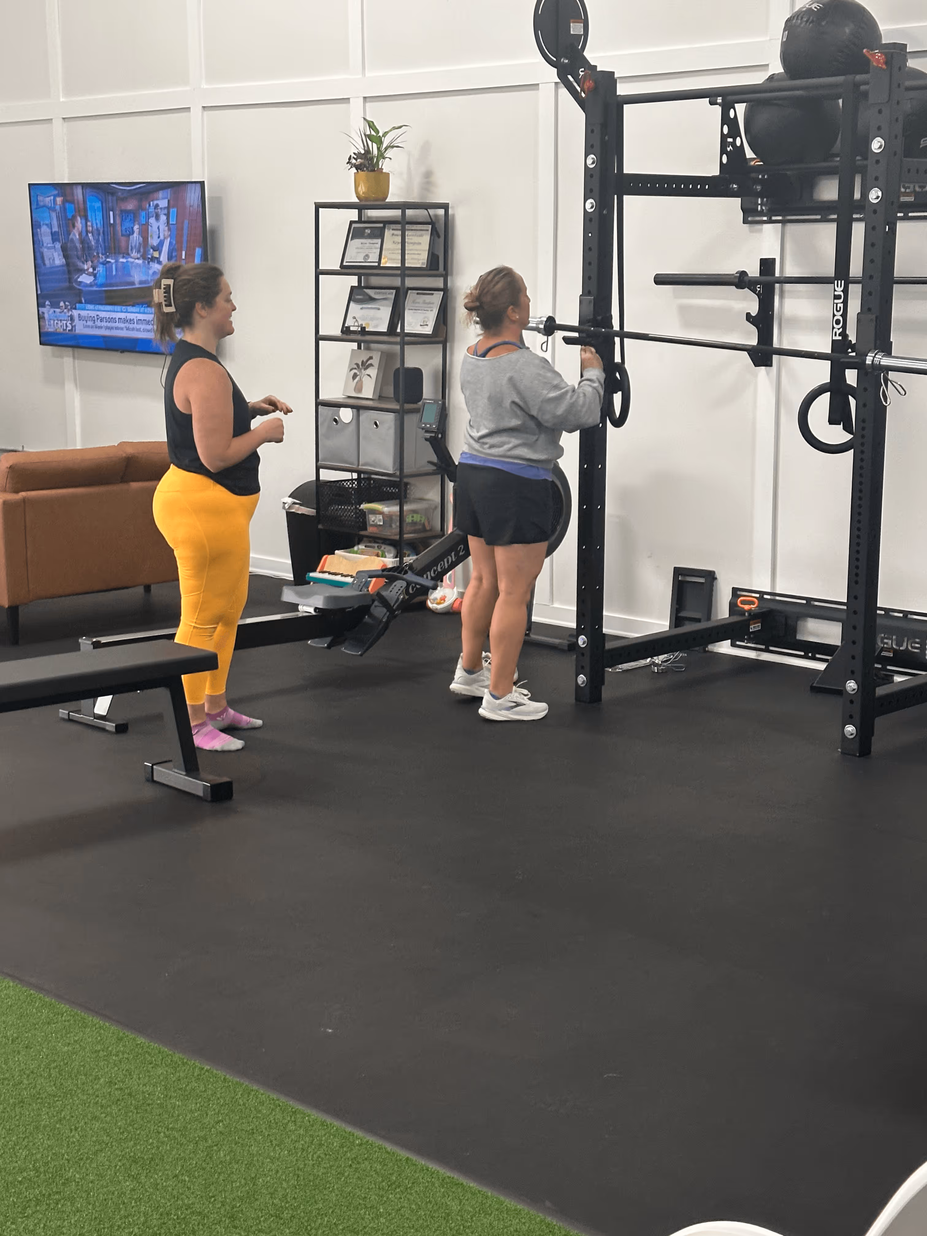 Two women exercising in a gym with weightlifting equipment and a rowing machine, with a TV and a bookshelf in the background.