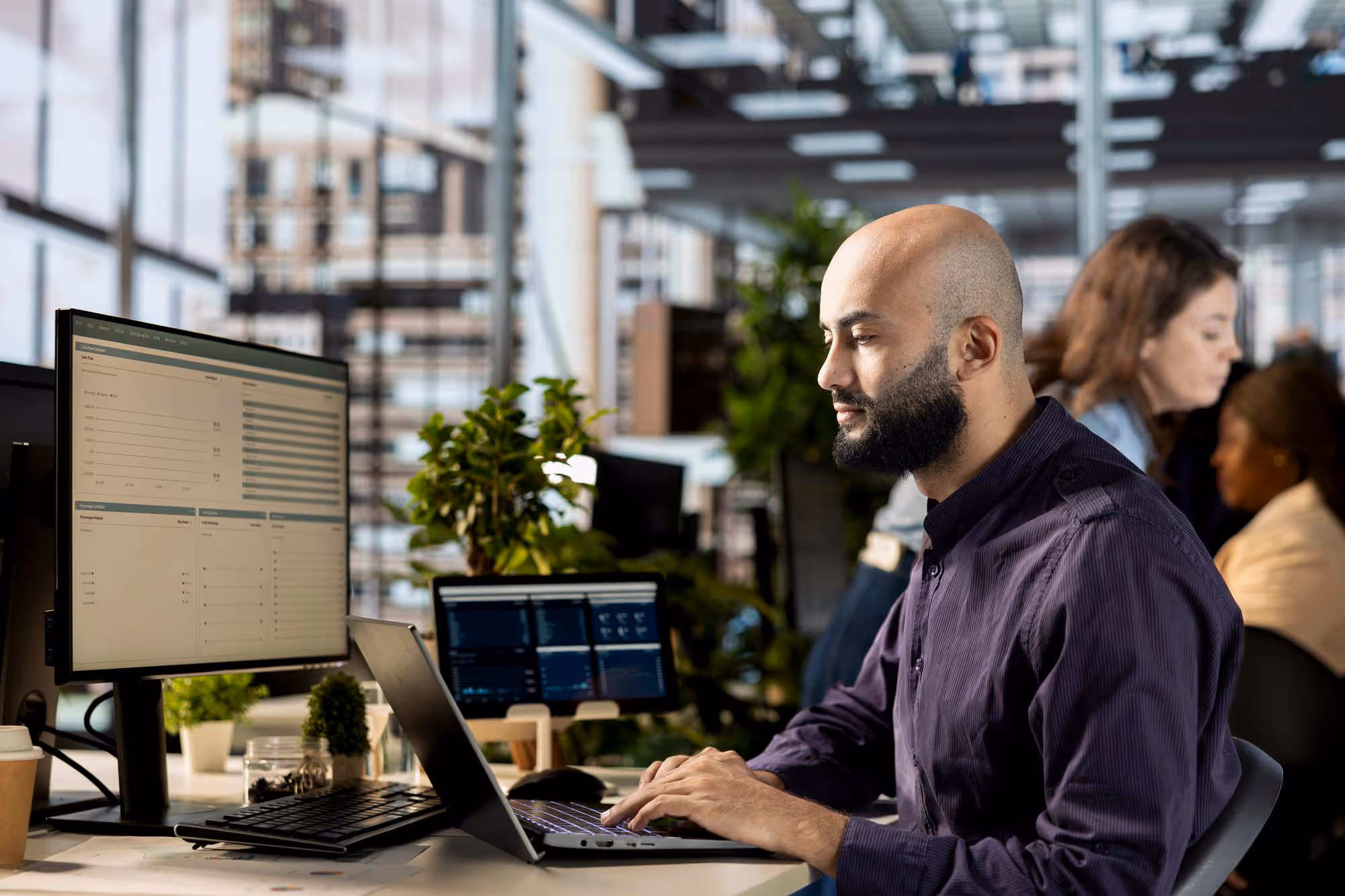 Man with beard working on a laptop at a desk with two monitors and plants in a modern office.