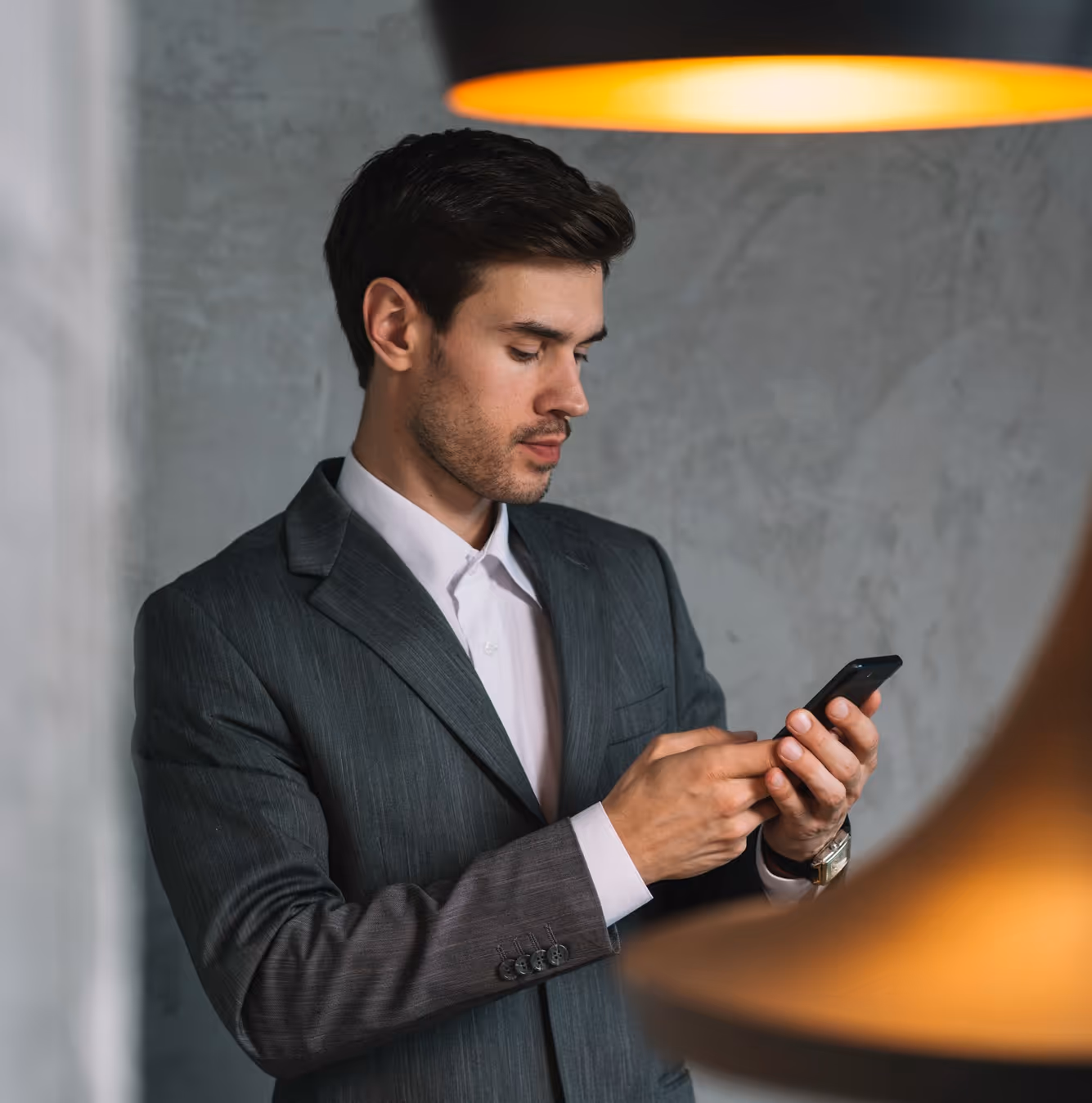 Man in gray suit looking at his phone with gray textured wall and orange pendant light in background.