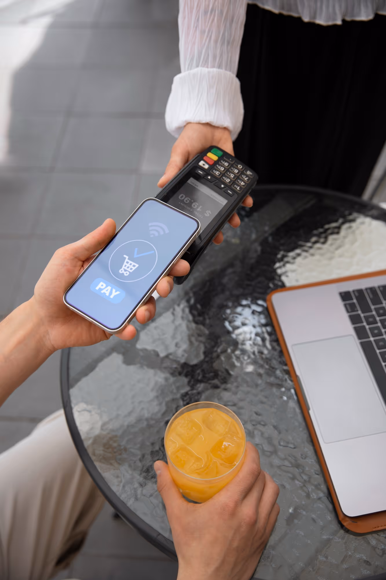 Person holding a smartphone with a digital pay screen near a card payment terminal, while the other hand holds a glass of orange juice on a glass table with a laptop.