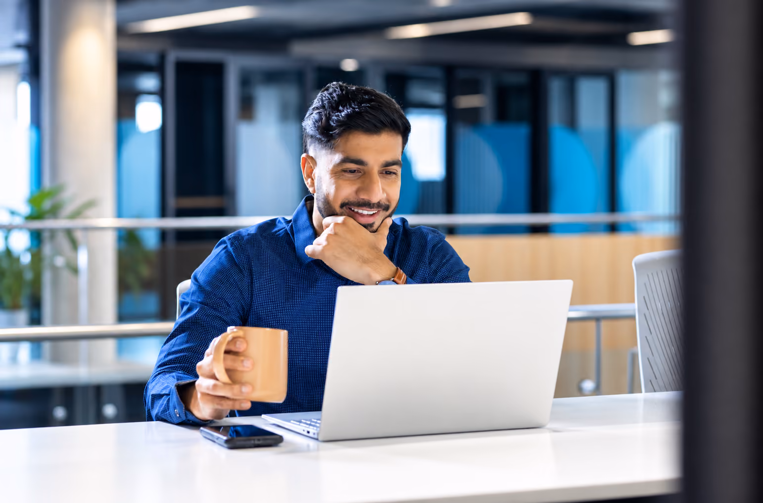 A person gazes at his laptop with a smile while holding a cup.