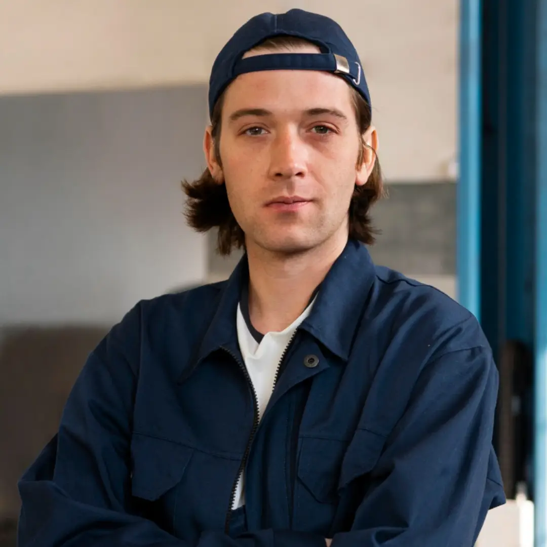 Young man wearing a backward navy cap and navy jacket with arms crossed, looking confidently at the camera indoors.