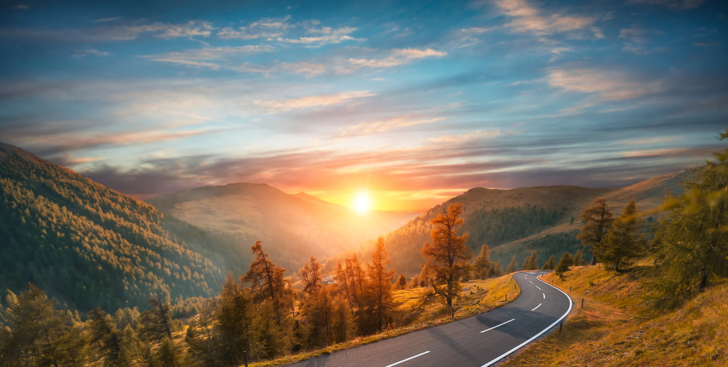 Gebirgsstraße bei Sonnenuntergang mit orangefarbenen Bäumen und bewaldeten Hügeln unter einem dramatischen Himmel.