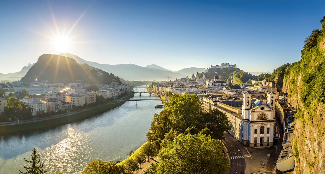 Panoramablick auf Salzburg mit der Salzach, historischen Gebäuden und der Festung Hohensalzburg im Sonnenschein.