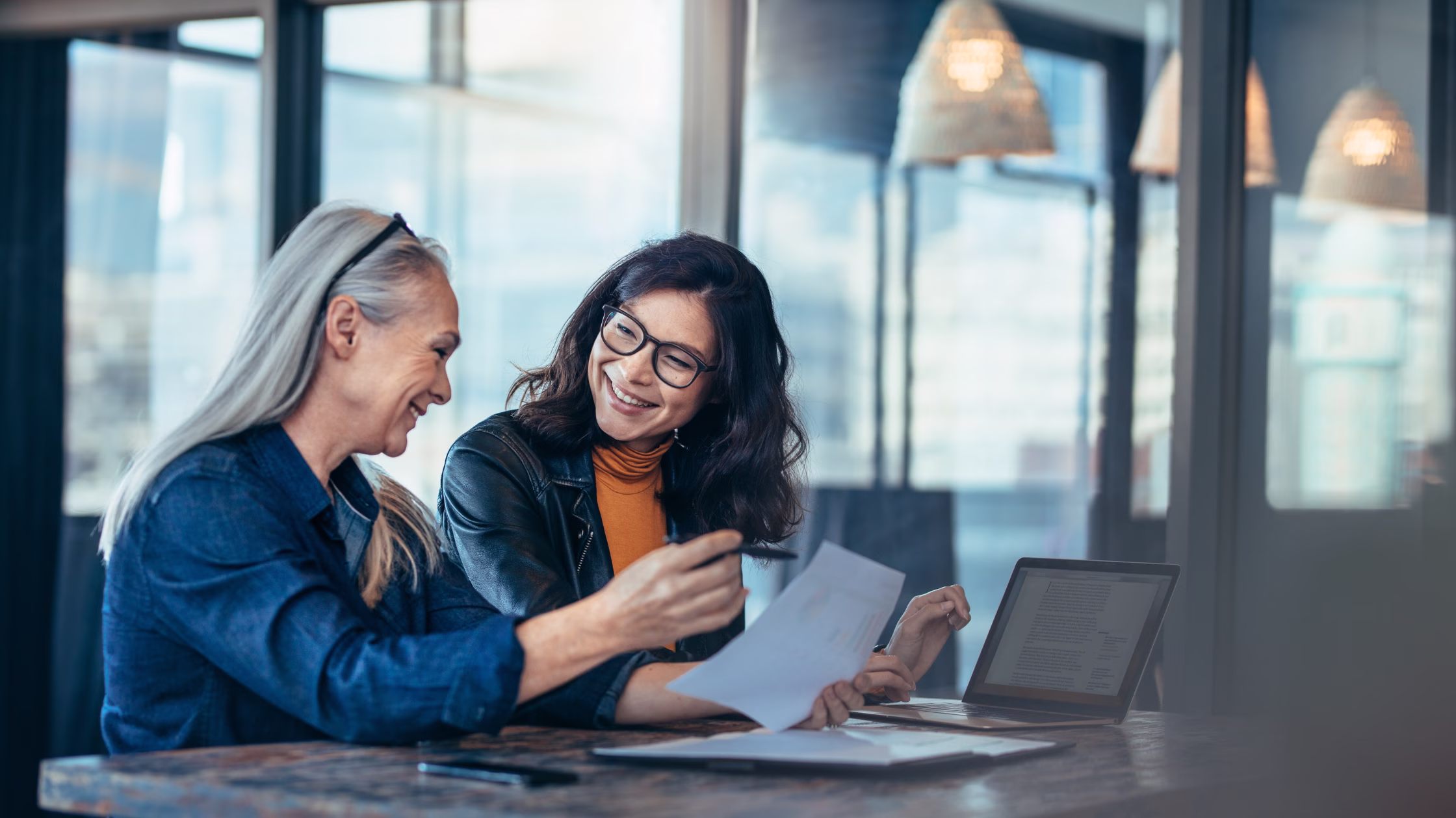 Two women review documents together at a coworking space, discussing membership options with a laptop and paperwork on the table.