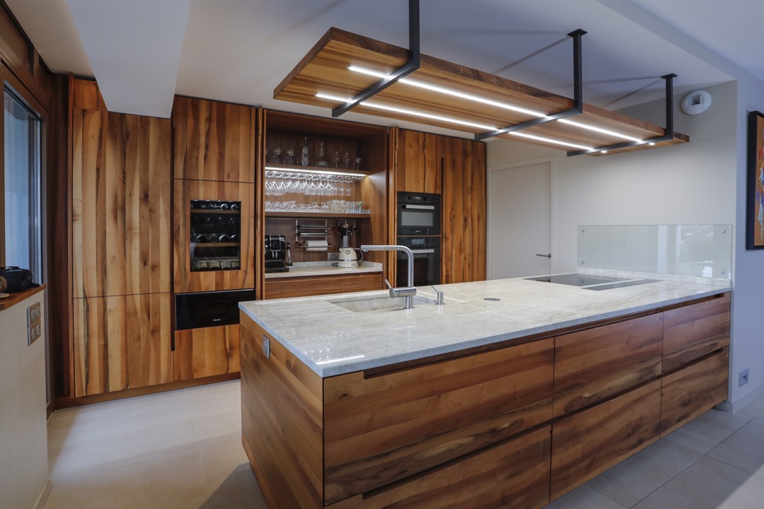 Moderne kjøkkenøy med marmortopp og trebunn, innebygd vask, oppvaskmaskin og veggskap i tre med integrerte ovner og vinlagring.