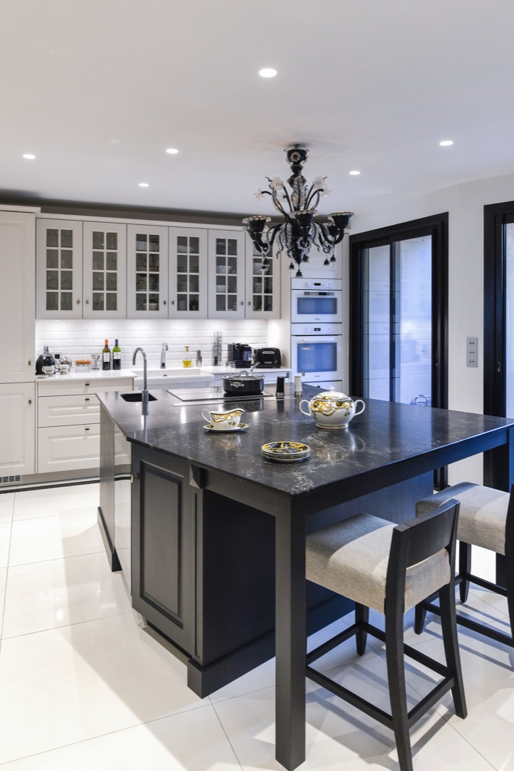 Moderne kjøkken med hvite skap, svart kjøkkenøy med servise, og en elegant svart lysekrone i taket.