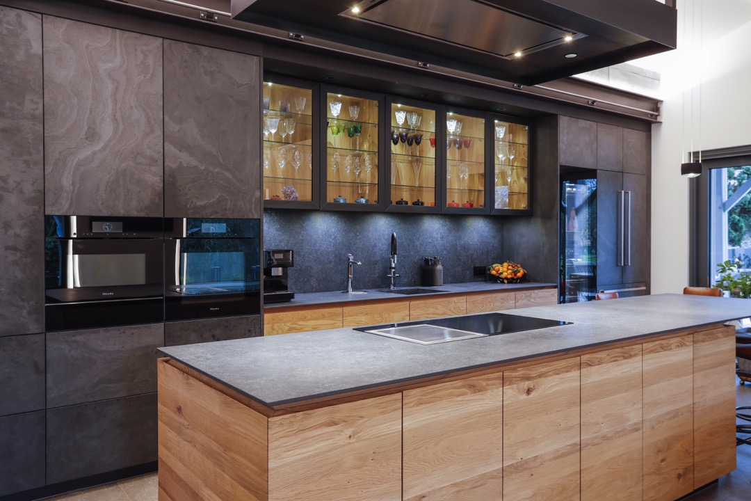 Moderne kjøkken med trekabinett, mørke flater, glassskap med glass og keramisk koketopp på øy.