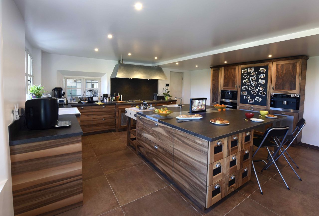 Moderne kjøkken med stor øy i midten, treskap, svart benkeplate og kjøkkenutstyr, samt to sammenleggbare stoler.