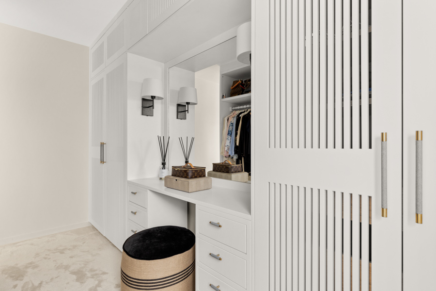 Moderne garderobeløsning i hvitt med integrert sminkebord, speil, skuffer og oppbevaring, med en rund puff foran.
