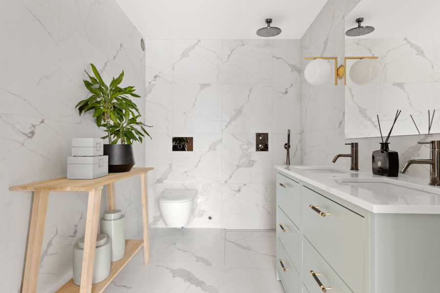Moderne baderom med hvite marmorfliser, vegghengt toalett, dobbel vask med gulldetaljer og et lyst tregulvskap med planter og dekorasjoner.