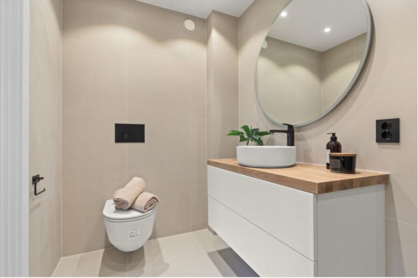 Moderne bad med vegghengt toalett, to foldede håndklær og en hvit servant på en treplate under et rundt speil.
