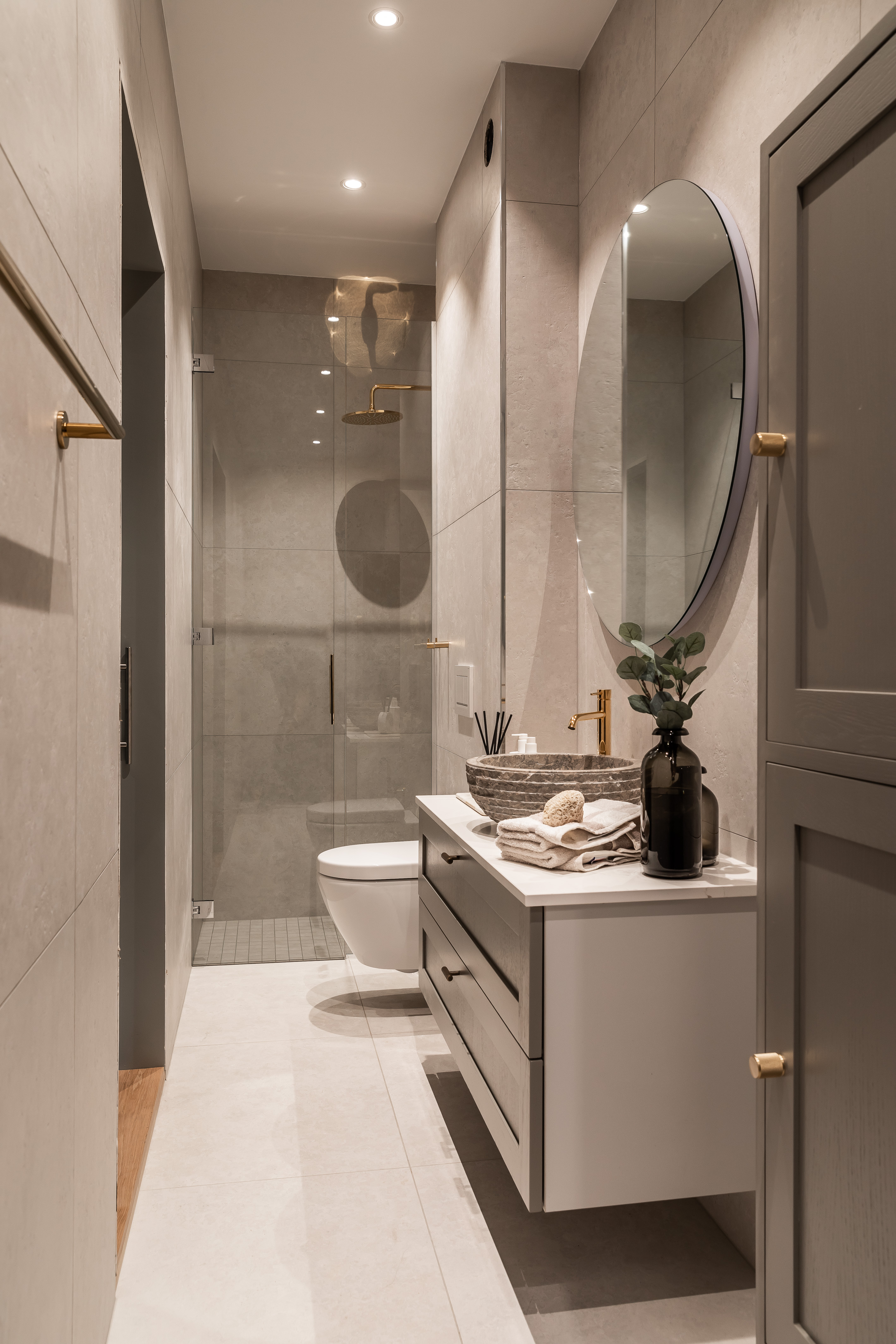 Moderne baderom med beige fliser, flytende toalett, glassdusj med gull dusjhode, rundt speil, vask med steinskål og hvit benk med håndklær og vase.