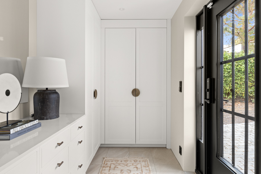 Lyst og moderne entré med hvite skap, mørk bordlampe, dekorativ skulptur og stor svart glassdør som fører ut til hage.