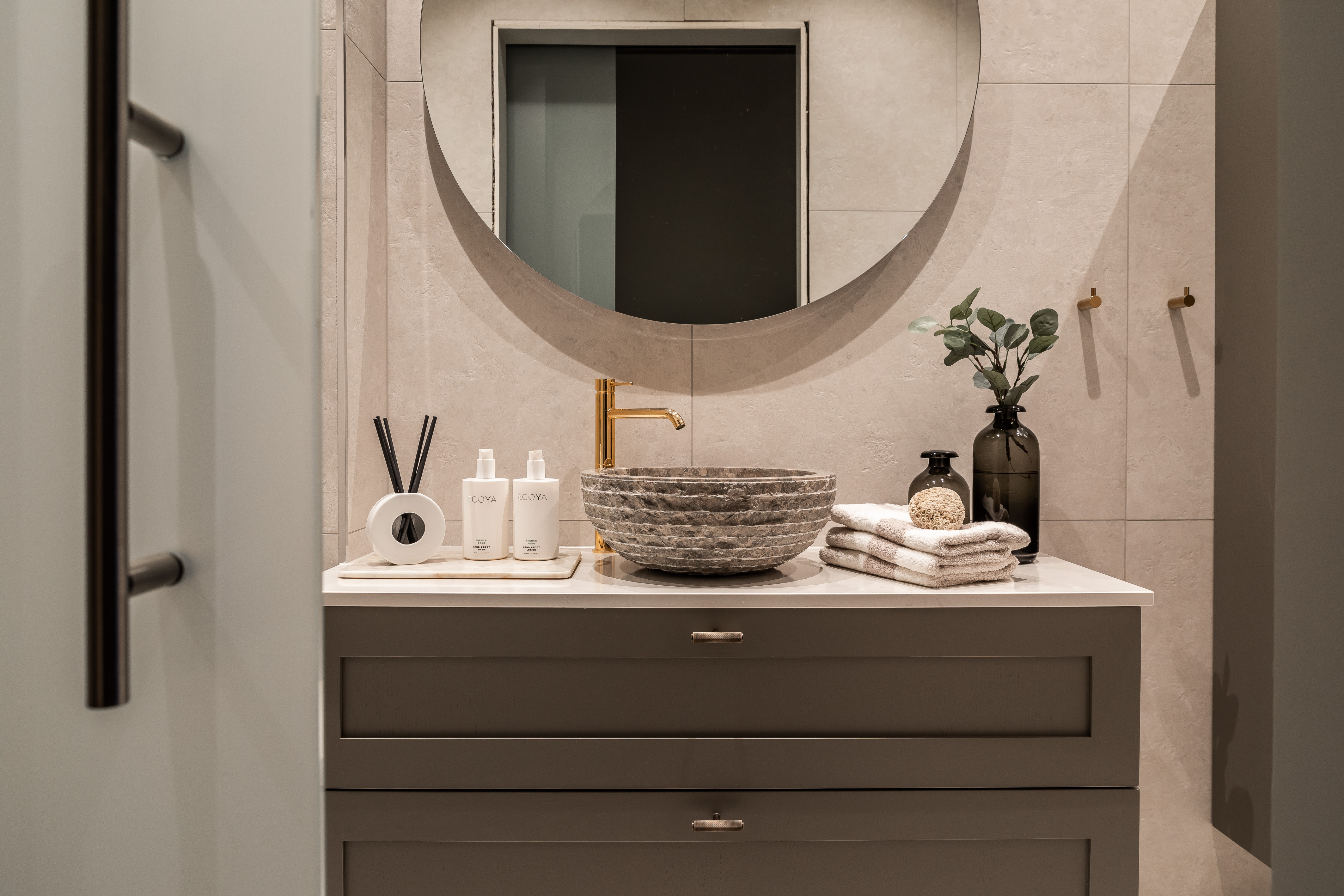 Moderne baderomsvask med steinskål, gyllen kran, sirkelrundt speil, håndklær og dekorative flasker på benkeplate.