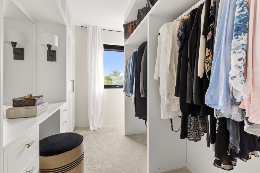 Lyst og ryddig klesrom med hvite hyller, hengende klær, en puff og vindu med hvite gardiner.