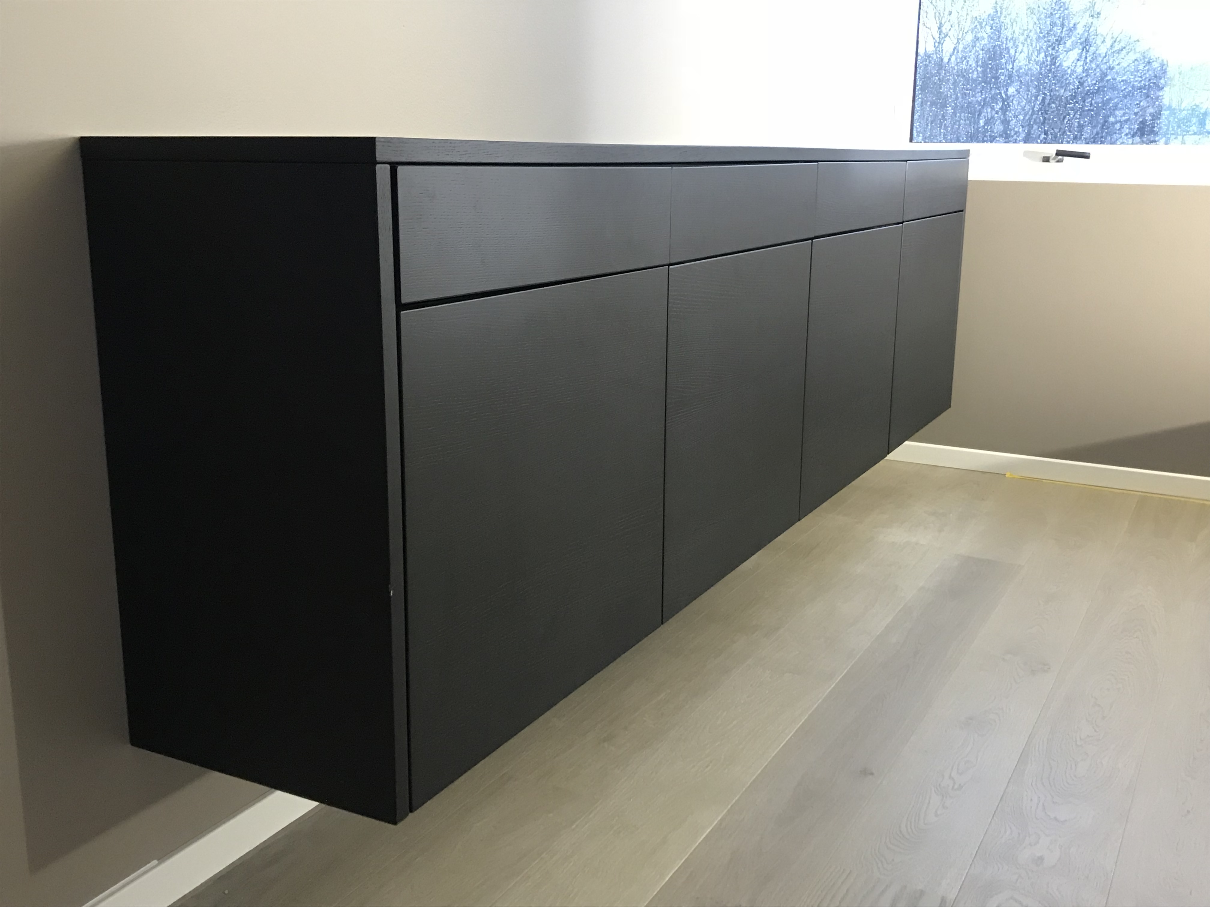 Sølvgrå vegghengt moderne svart skap med dører og skuffer i lyst rom med parkettgulv og vindu.