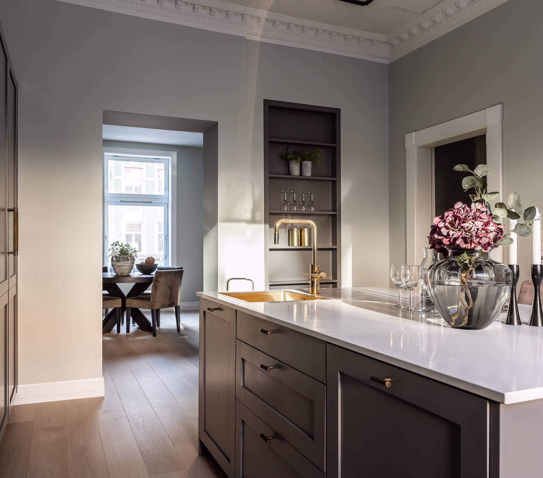 Moderne kjøkkenøy med hvit benkeplate, gyllen kjøkkenkran og glassvase med rosa blomster, med utsikt til spisebord i bakgrunnen.