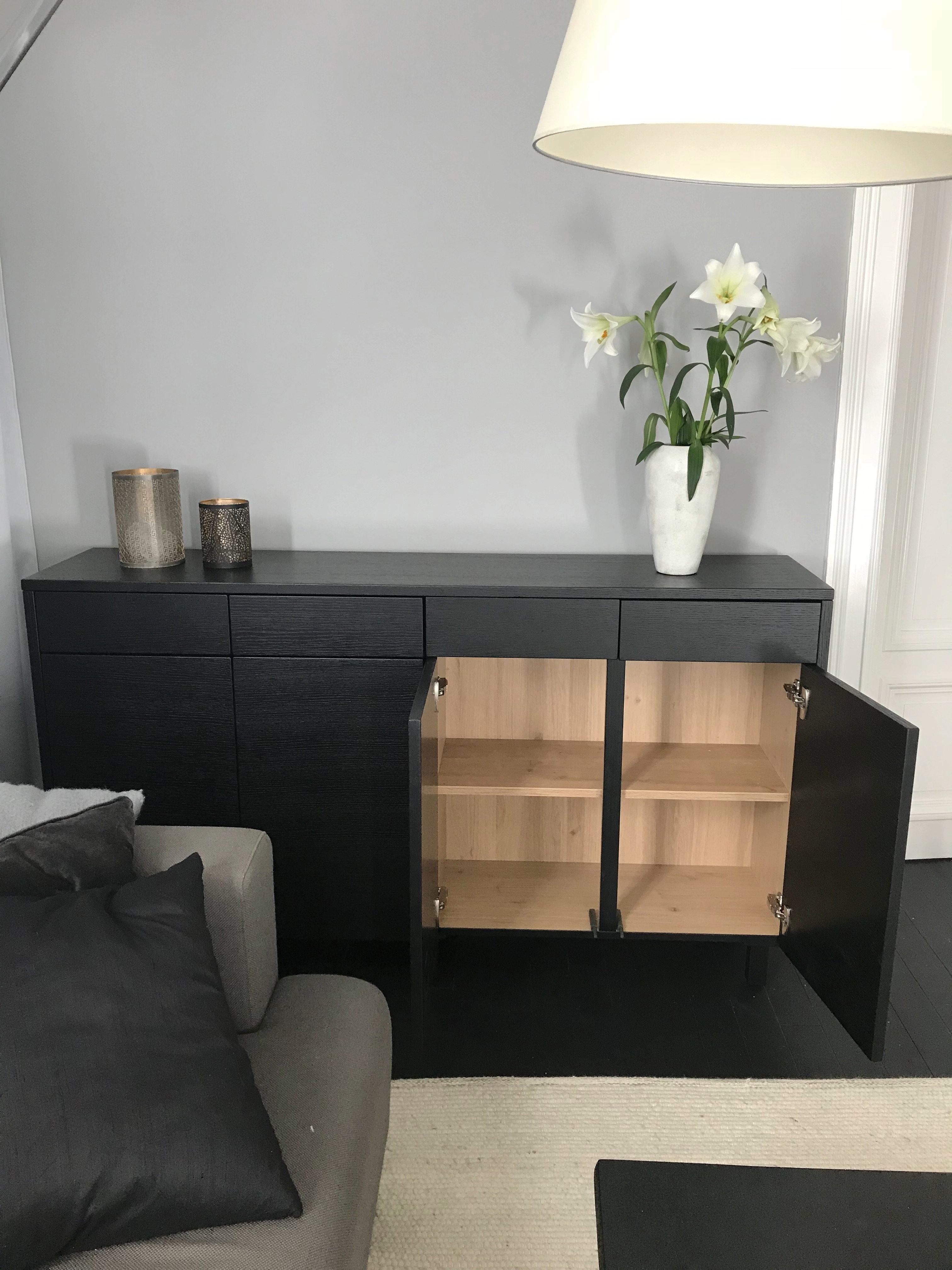 Svarte skap med to åpne dører som viser hyller inni, med hvite blomster i en vase og to lykter på toppen, ved siden av en grå sofa med svarte puter.