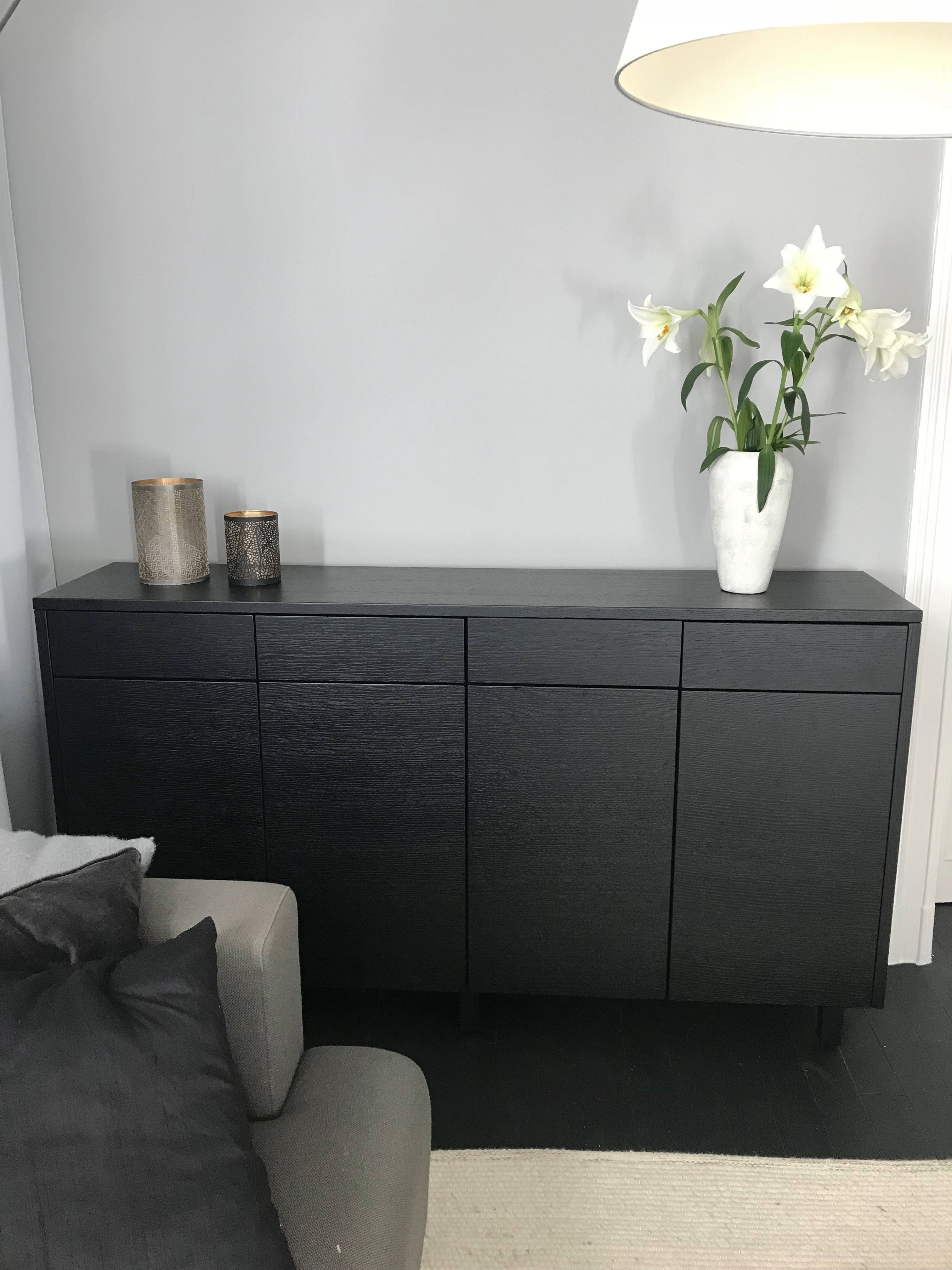 Svart kommode med to dekorative lysestaker og en hvit vase med hvite liljer, ved siden av en grå sofa med puter.