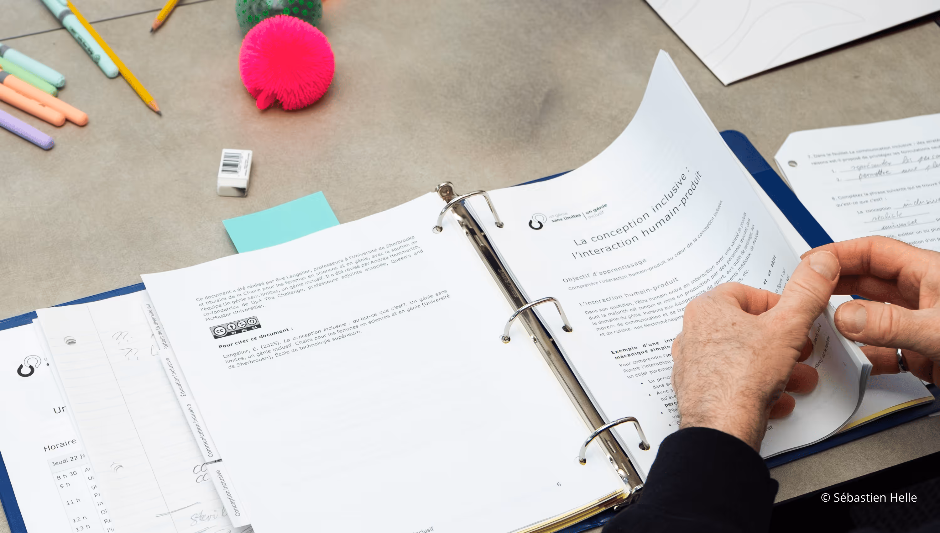 Une personne tourne les pages d'un classeur contenant des documents imprimés en français, posés sur une table avec des crayons, gommes et objets colorés.