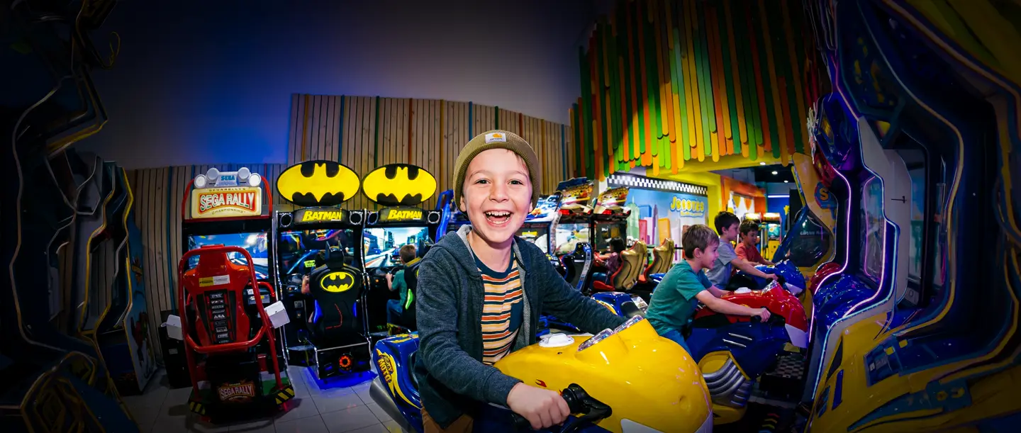 Niño sonriente con un gorro montado en una moto amarilla de arcade, con otros niños jugando juegos de carreras en el fondo.