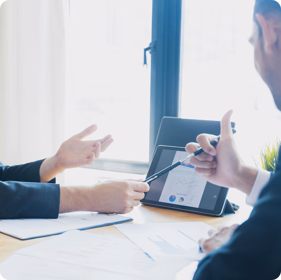 Deux personnes en costume discutent autour d'une tablette affichant des graphiques et des documents sur une table.