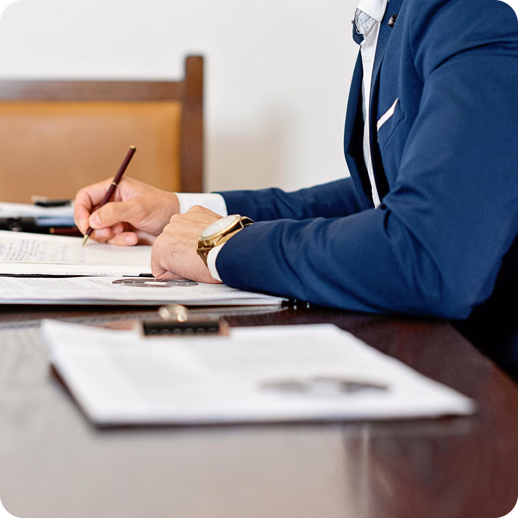 Une personne en costume bleu écrit sur un document avec un stylo, une montre dorée au poignet visible.