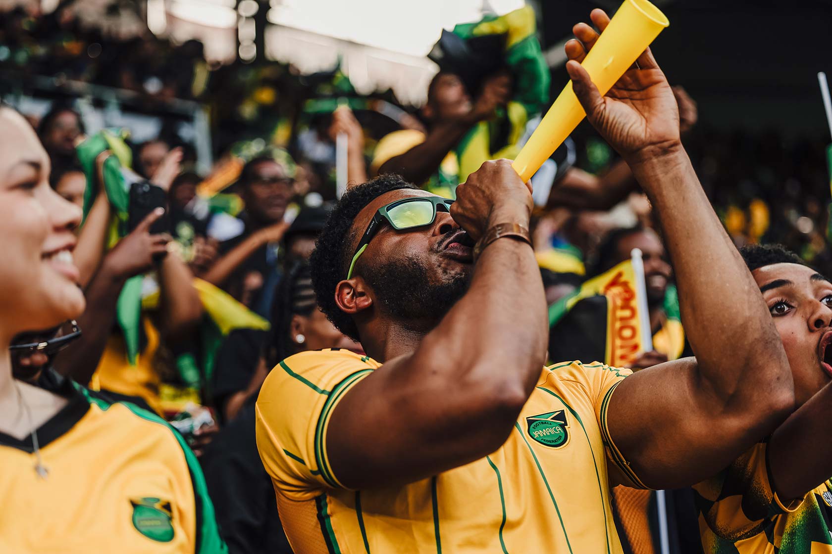 Crowd of Jamaican soccer fans cheering, one blowing a yellow vuvuzela horn.