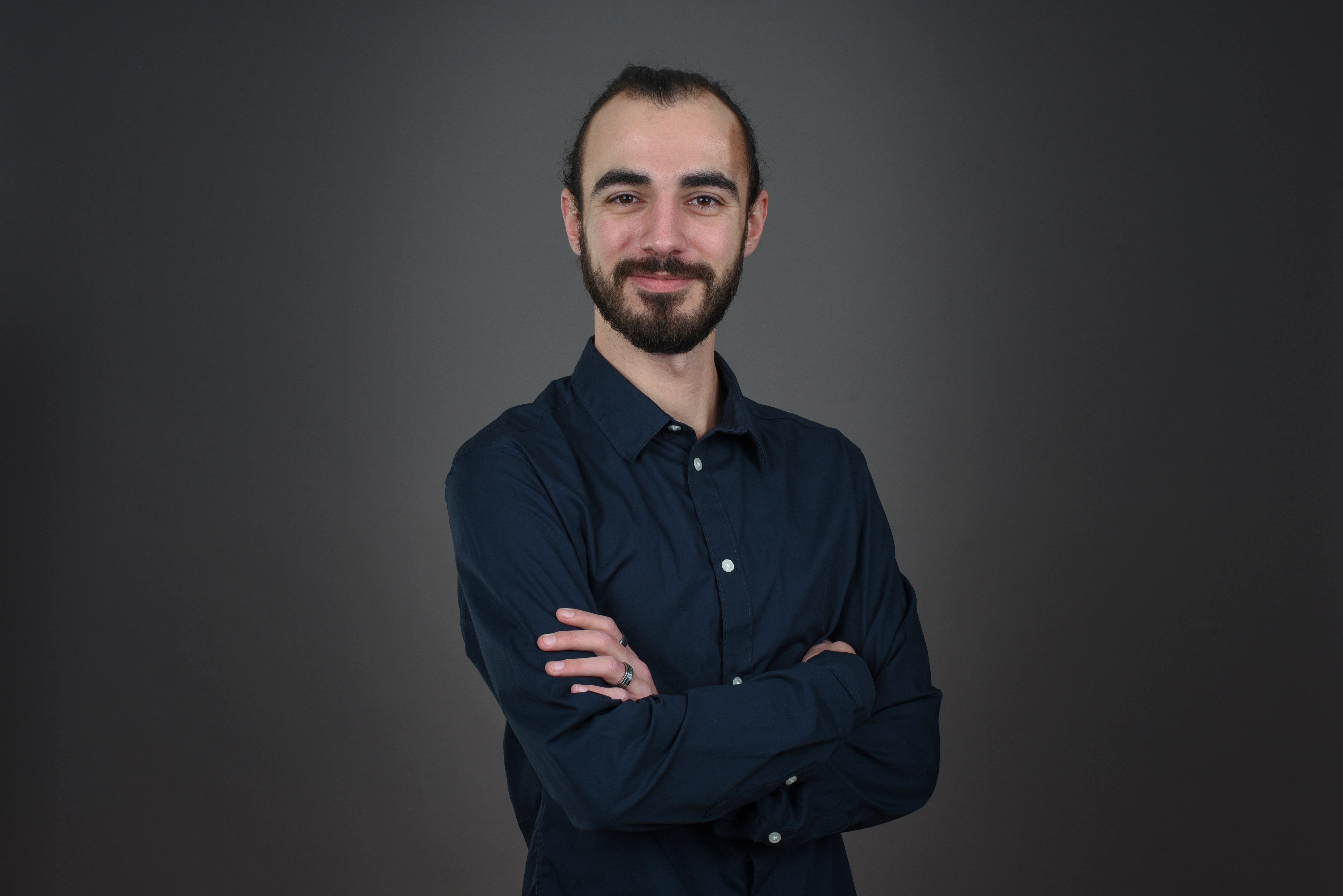 Smiling man with a beard and mustache wearing a dark blue button-up shirt standing with arms crossed against a gray background.