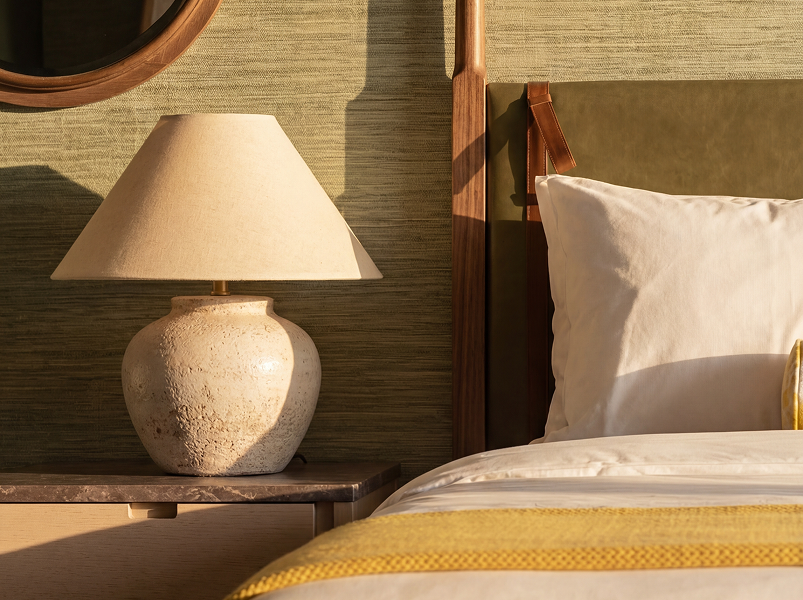 Beige textured table lamp on a bedside table next to a bed with white pillow and yellow blanket.
