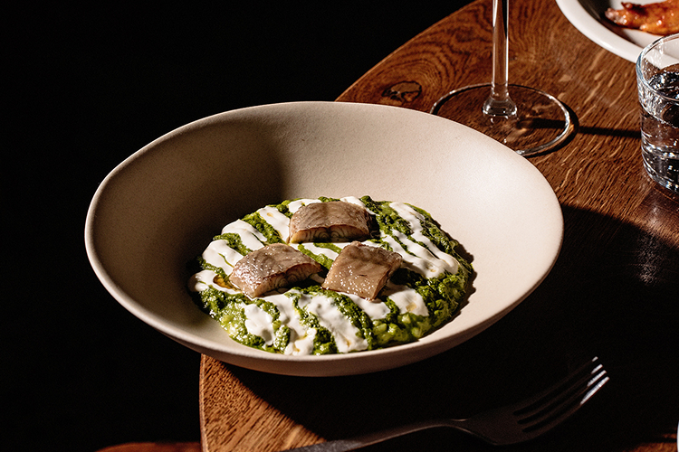 Beige bowl with green creamy dish topped with white sauce and three pieces of cooked fish, on a wooden table next to a wine glass and a fork.