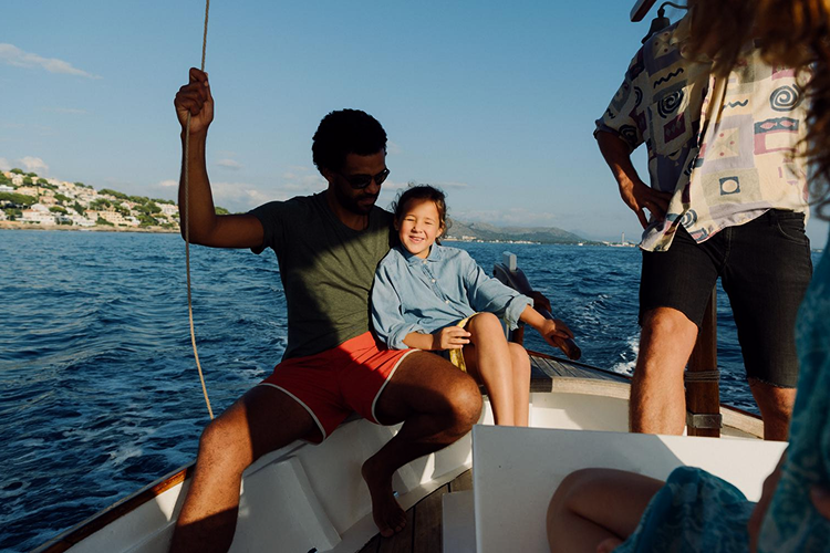 A man wearing sunglasses and red shorts holding a rope on a boat with a smiling young girl sitting beside him and two other people partially visible.