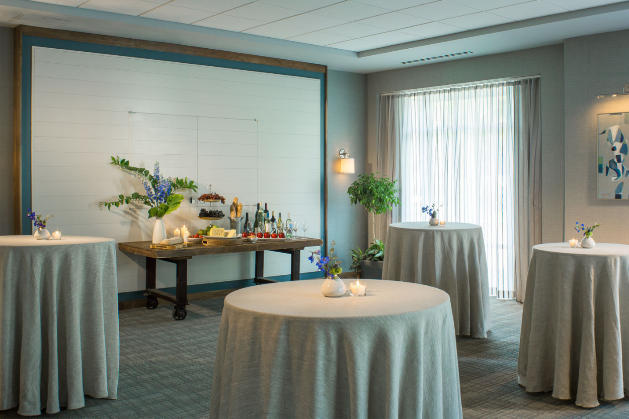 The Independence Room at the Harborvale set for a reception with round tables covered in beige cloths, small vases with purple flowers, lit candles, and a wooden table with wine bottles, glasses, and snacks.