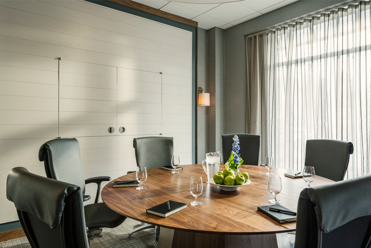 The Enterprise Room at the Harborvale set up as a conference room with a round wooden table, six black leather chairs, notebooks, wine glasses, a water pitcher, and a bowl of green apples with a purple flower centerpiece, next to large windows with sheer curtains.