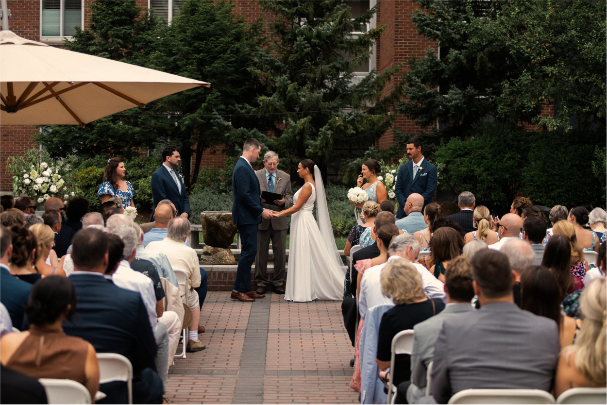 An outdoor wedding ceremony with bride and groom holding hands in front of officiant surrounded by wedding party and seated guests on the terrace of the Harborvale in Burlington, Vermont.