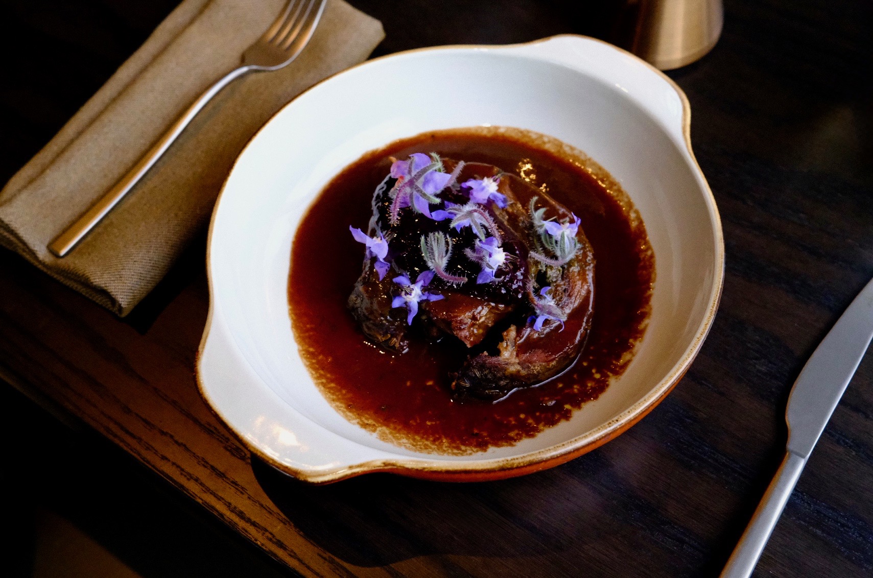 Dark meat dish in brown sauce garnished with small purple edible flowers on a white plate.