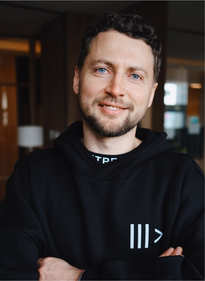 Smiling man with blue eyes and light beard wearing a black hoodie with a white symbol, standing indoors with arms crossed.