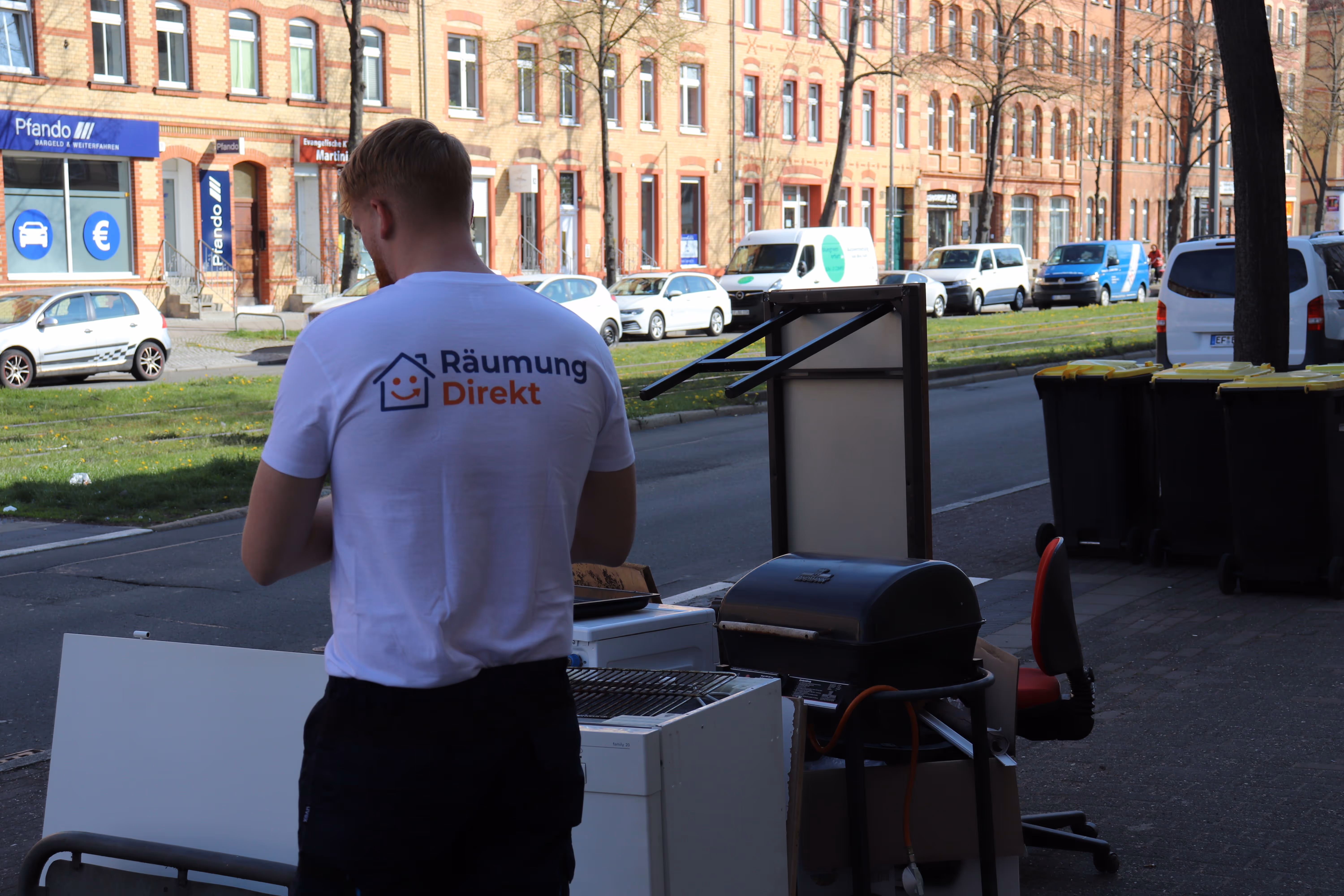 Mitarbeiter mit einem weißen T-Shirt mit der Aufschrift 'Räumung Direkt' steht neben Sperrmüll auf einem Bürgersteig in einer Straße mit parkenden Autos.
