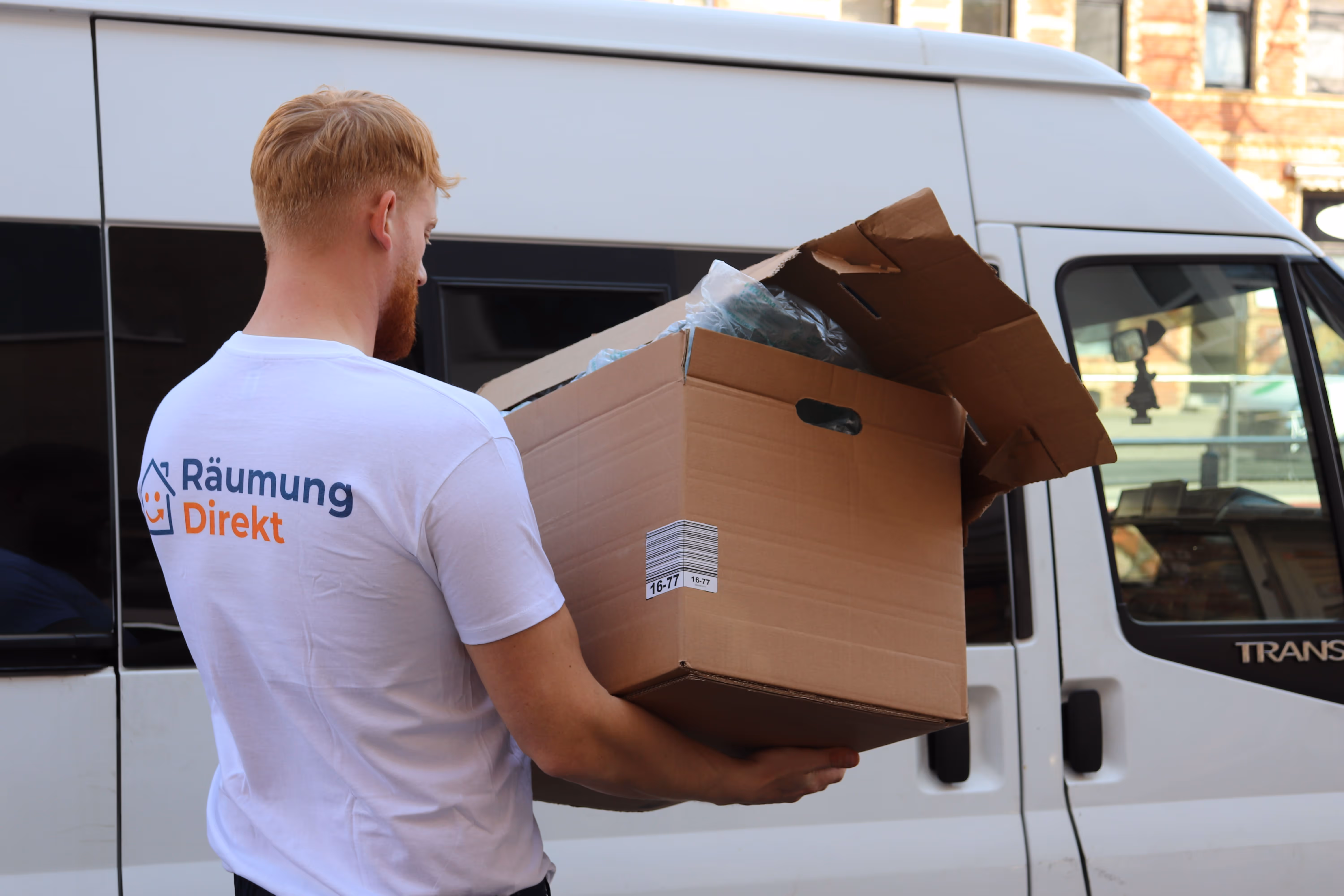 Mitarbeiter mit weißem T-Shirt mit dem Logo Räumung Direkt trägt einen geöffneten Karton vor einem weißen Transporter.