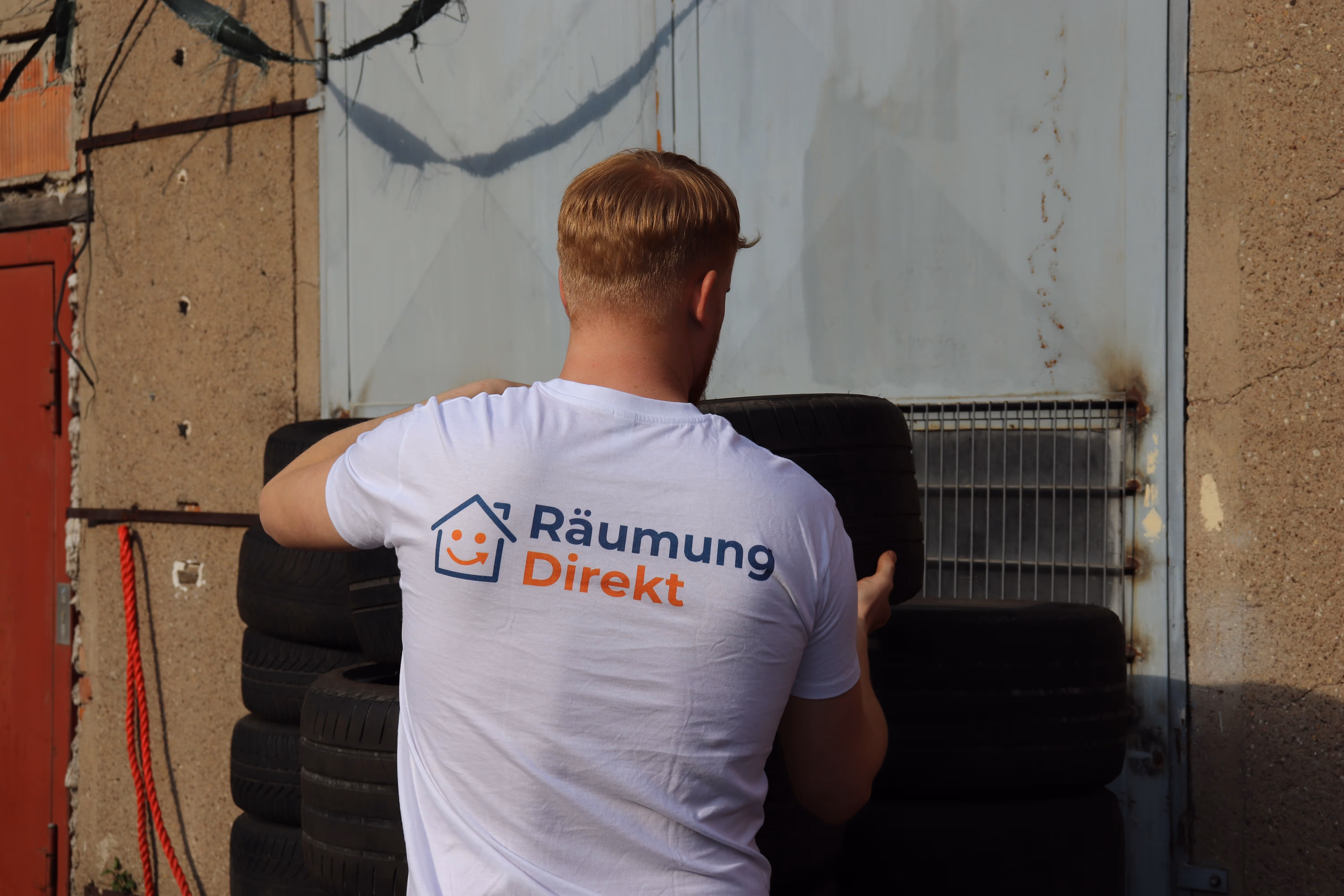 Mitarbeiter mit weißem T-Shirt mit Aufschrift 'Räumung Direkt', der Autoreifen vor einer Garage stapelt.