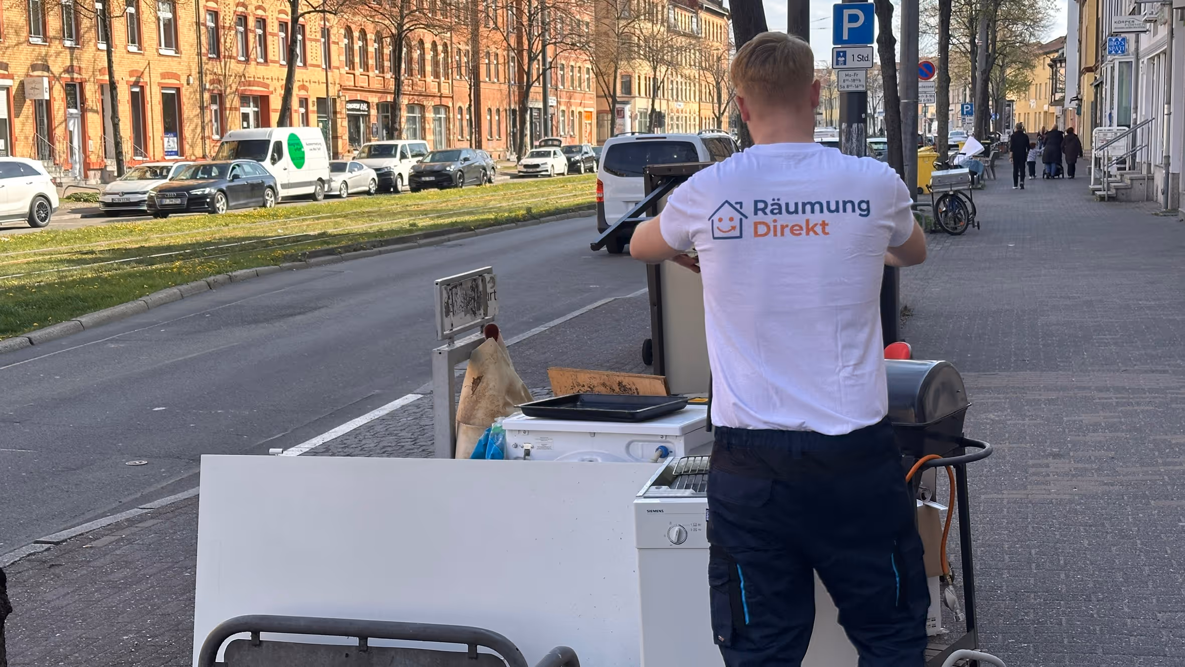 Mitarbeiter mit weißem T-Shirt mit Aufschrift 'Räumung Direkt' schiebt Haushaltsgeräte und Möbel auf einem Bürgersteig in einer Stadt.