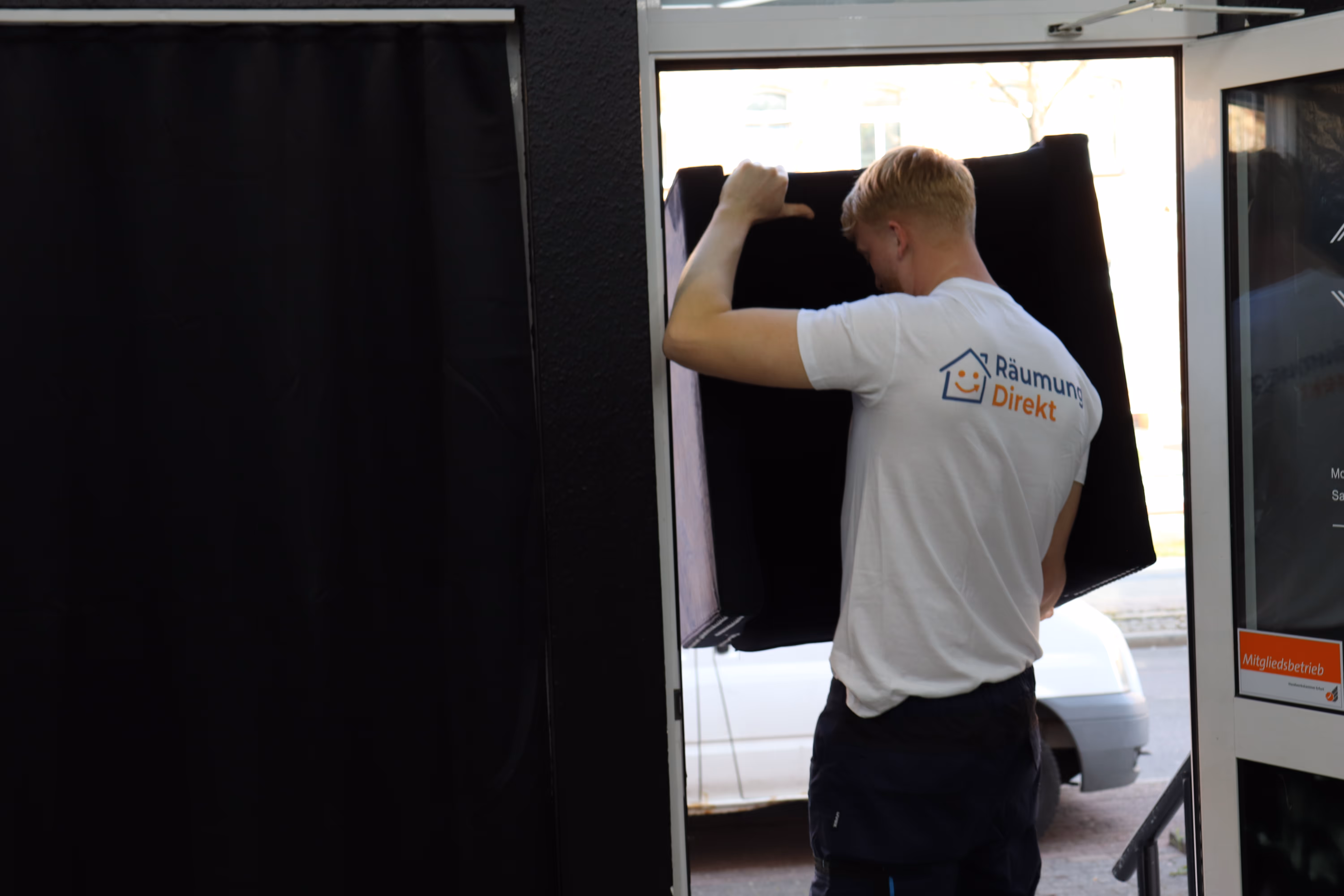 Mitarbeiter trägt schwarzen gepolsterten Gegenstand durch eine offene Tür und trägt ein weißes T-Shirt mit Aufschrift ‚Räumung Direkt‘.