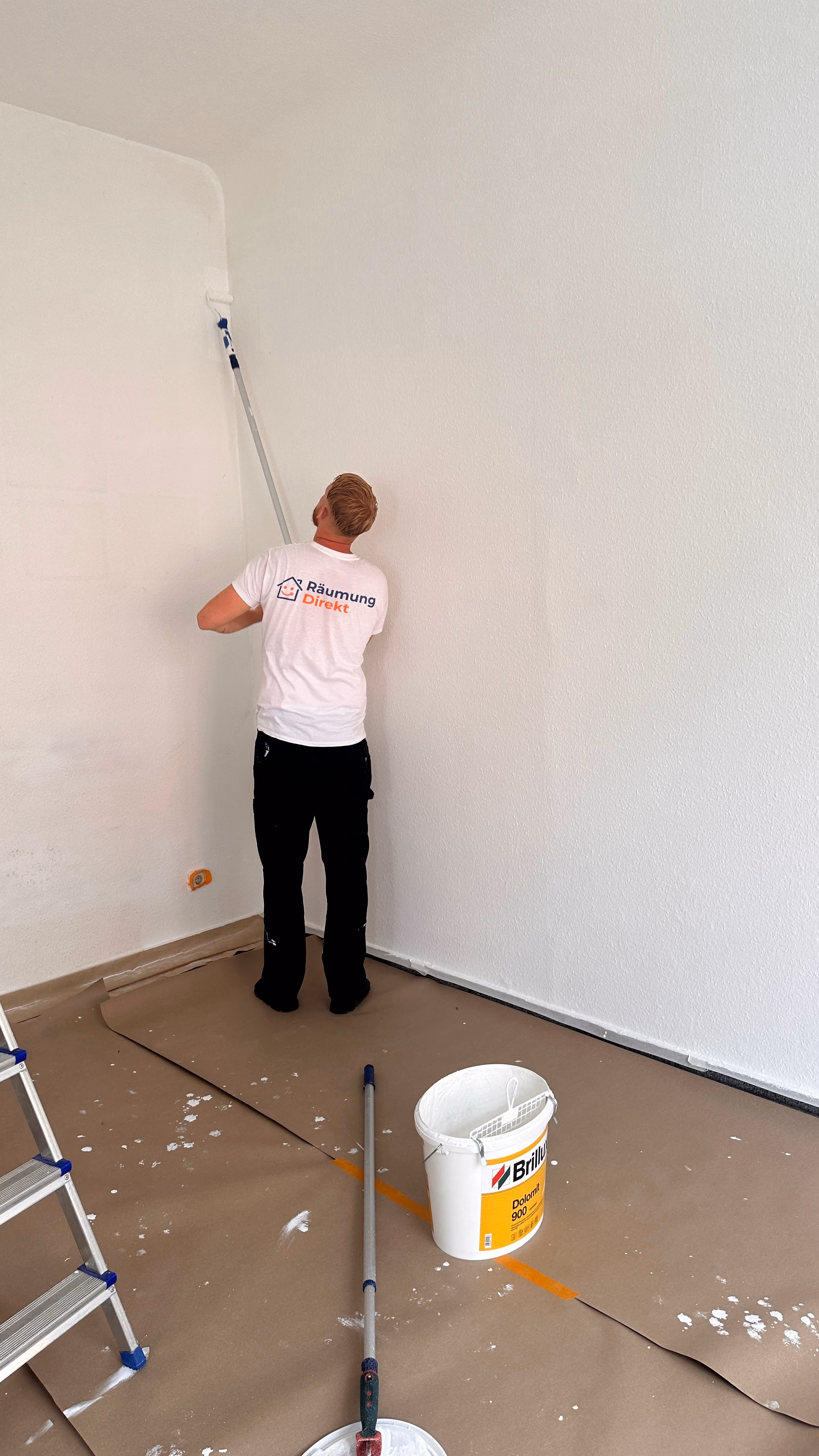 Mitarbeiter mit weißem T-Shirt streicht eine Wand mit Farbroller, Boden mit Papier abgedeckt.