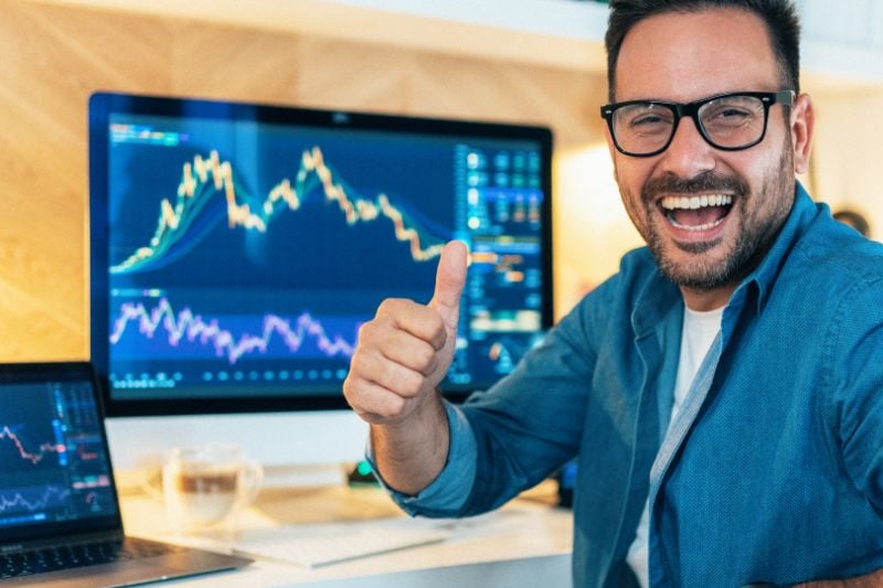 Smiling trader giving a thumbs-up in front of multiple trading screens displaying charts, symbolising success in completing a prop firm challenge.