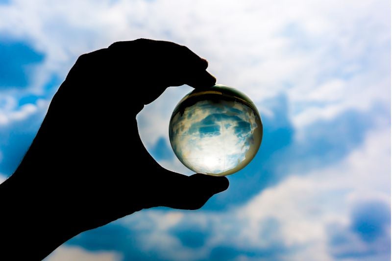 A hand holding a clear sphere reflecting the sky, representing the complexities of how prop firms generate profits.