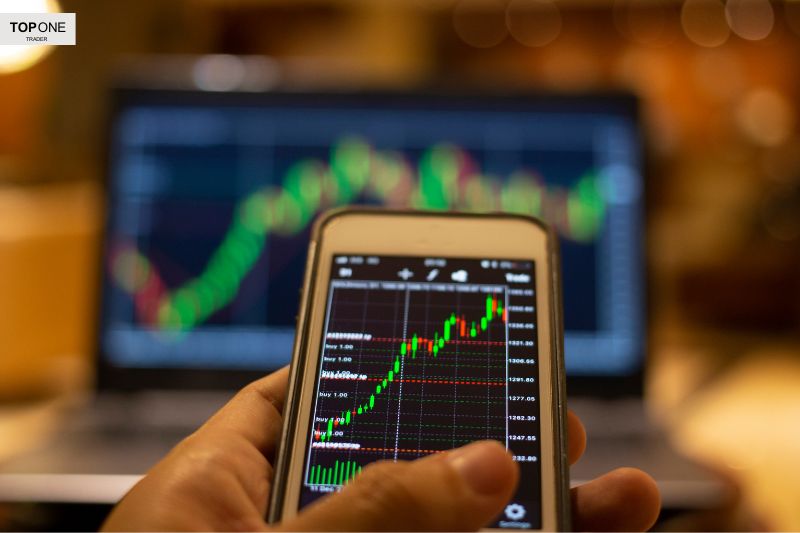 A trader analyzing forex charts on a smartphone with a laptop screen displaying market trends in the background