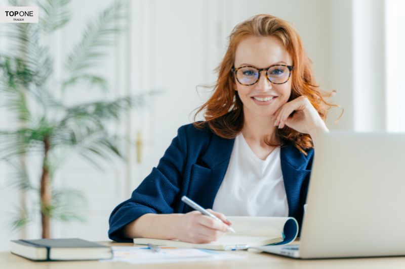 Smiling woman taking notes at her laptop, illustrating what is a funded trader and the skills needed for trading success
